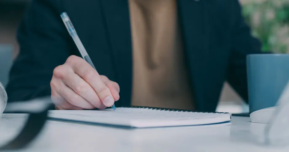 A person at a desk writing notes in a notebook beside a phone showing a suspicious text message